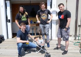 Four male students stand on the dock at the MIT Sailing Pavilion with a surface vehicle resembling a miniature boat that they developed