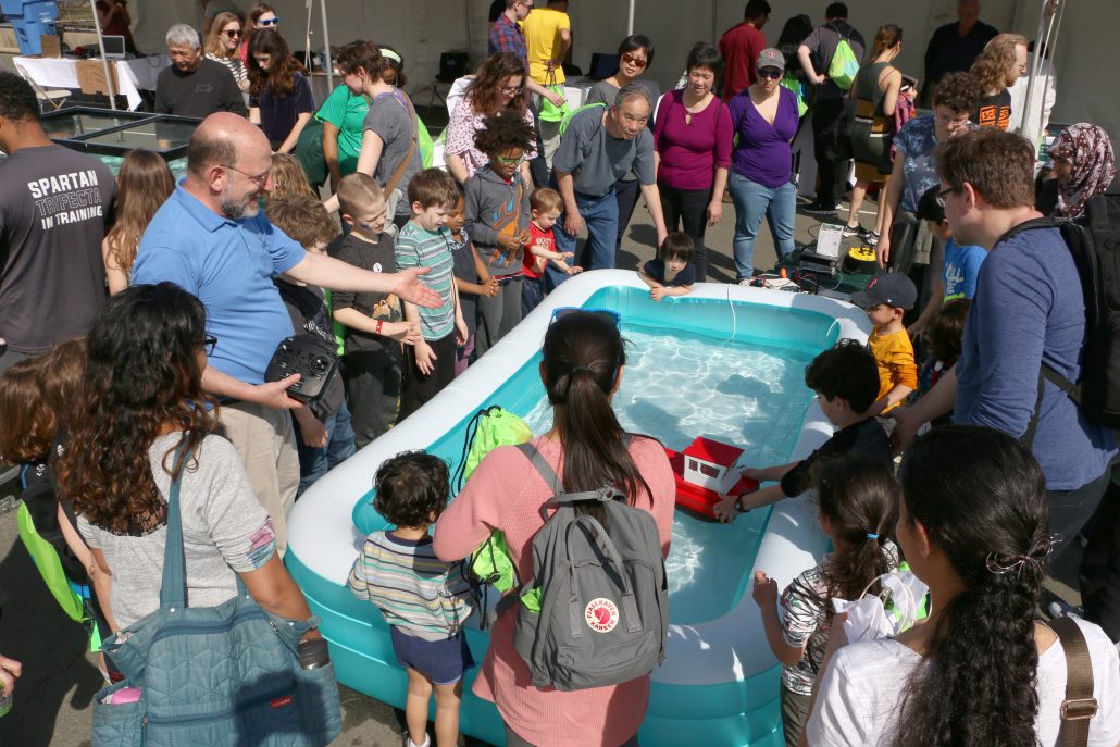 A large crowd of children and parents gather around the robotic fish pool for a demonstration