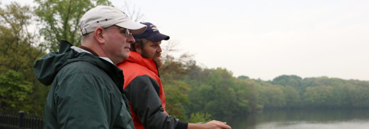 Two representatives from MIT Sea Grant and the MA DMF wearing baseball caps and rain gear look out over the Mystic Lakes