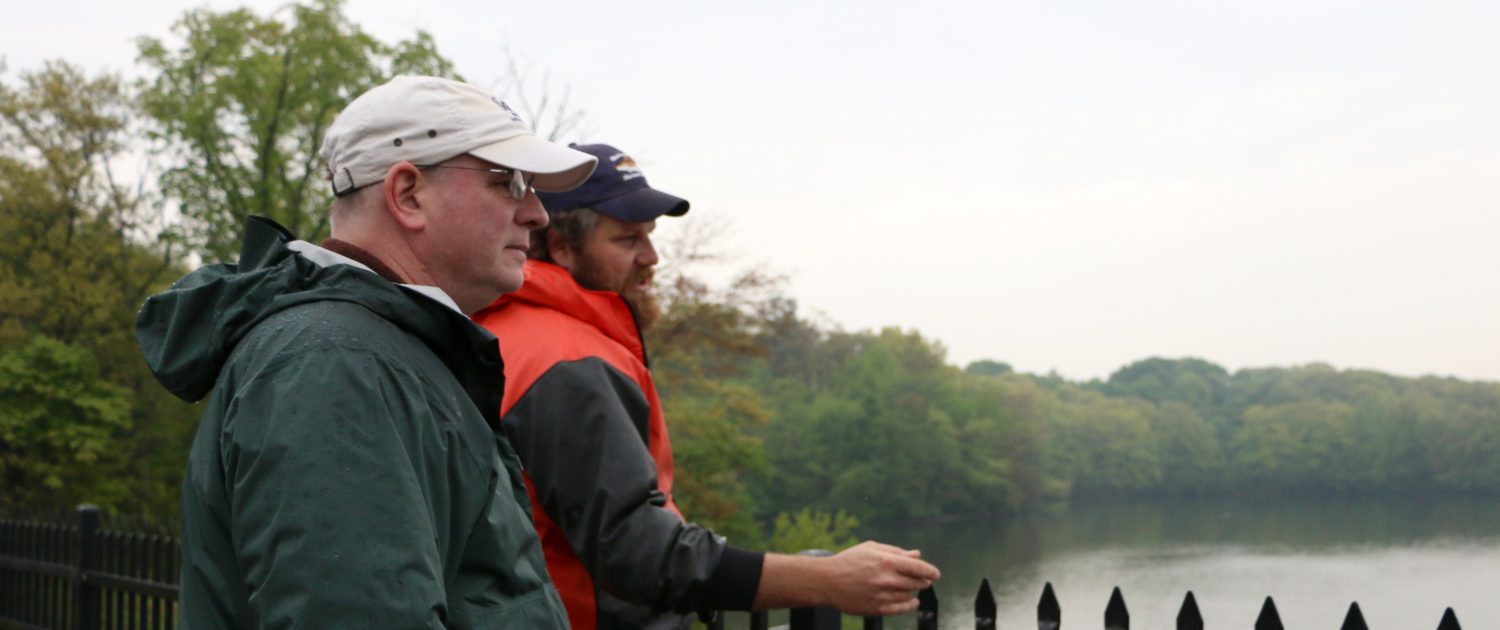 Two representatives from MIT Sea Grant and the MA DMF wearing baseball caps and rain gear look out over the Mystic Lakes