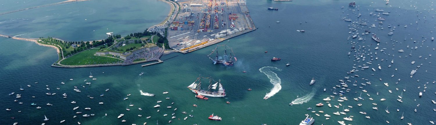 An aerial shot of a blue-green Massachusetts Bay and Castle Island with many boats and a couple of tall ships