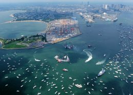 An aerial shot of a blue-green Massachusetts Bay and Castle Island with many boats and a couple of tall ships