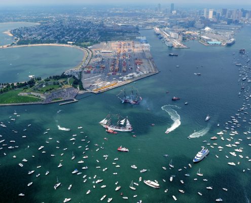 An aerial shot of a blue-green Massachusetts Bay and Castle Island with many boats and a couple of tall ships