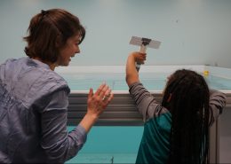 A woman and a child from behind about to drop a cardboard model into a large tank of water.