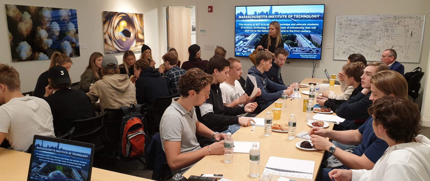 A group of students sitting for a presentation with a female speaker in front of a screen displaying a picture of the MIT campus