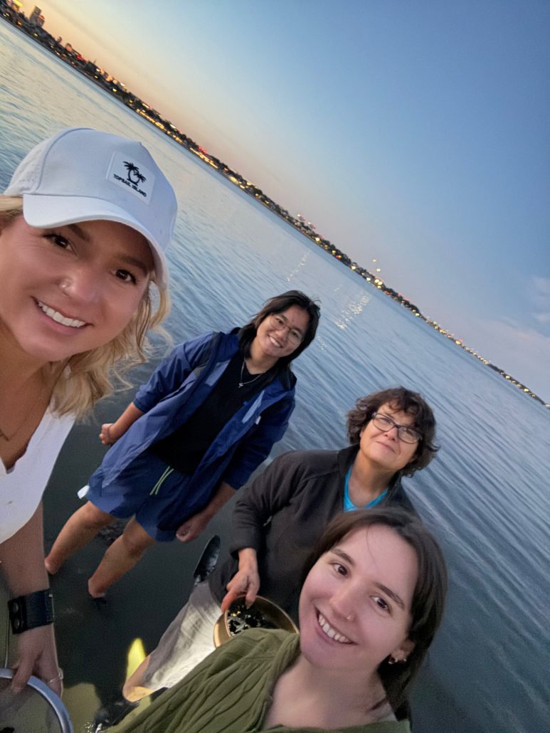 Scientists and students Aly Putnam, Isabella Yeung, Carolina Bastidas, and Addy Dwyer on a shore at sunset with Boston skyline in the background.